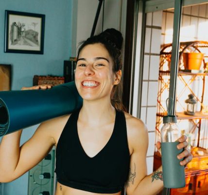 Cheerful woman with yoga mat and bottle ready for home workout in cozy room.