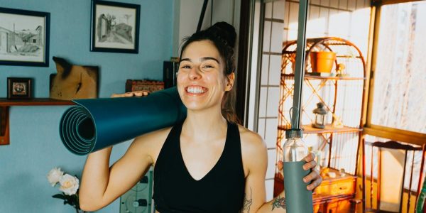Cheerful woman with yoga mat and bottle ready for home workout in cozy room.